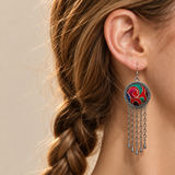 Scarlet Affinity earring worn at the ear, showing red, teal and magenta embroidery with pale curving outlines and five long chain tassels.