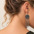 Model looking down in an olive sleeveless top, wearing the floral metal reverse of a Rose Chorus earring with five long tassel chains.