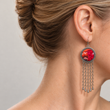 Ember Refrain earring worn at the ear, showing raised red and orange embroidery on pale pink, a patterned rim and five long tassel chains.