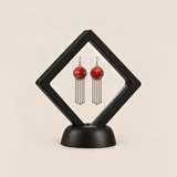 Ember Refrain earrings displayed in a tilted black square membrane frame on a round black base.