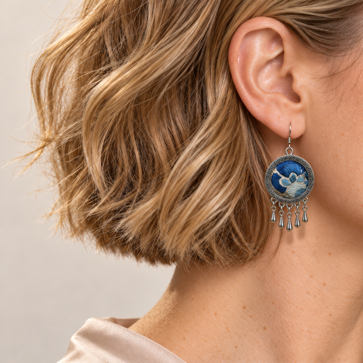 Azure Harmony earring worn at the ear, showing blue floral embroidery on ivory-colored cloth, a patterned rim and five short teardrops.